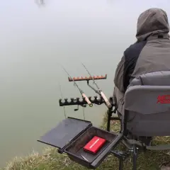 Wędkarz siedzi nad wodą, gotowy do łowienia karpia. Przed nim rozłożony jest **zestaw na karpia metodą** feeder, z wędkami i pudełkiem na przynęty.