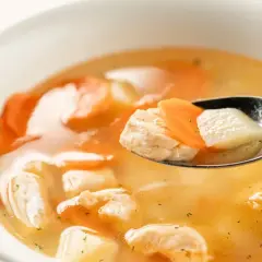 Łyżka z kawałkami kurczaka i marchewki zanurzona w aromatycznej zupie rybnej.
