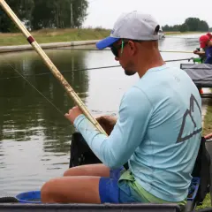 Mężczyzna w czapce i okularach przeciwsłonecznych siedzi nad wodą, skupiony na łowieniu na spławik. W tle widać innych wędkarzy.