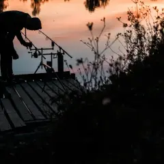 Wędkarz na pomoście przygotowuje sprzęt. W tle zachód słońca nad wodą, idealne warunki na leszcza.