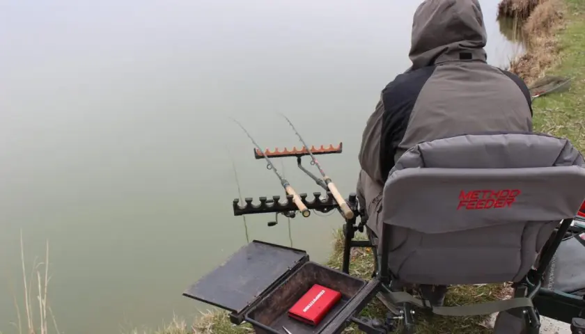 Wędkarz siedzi nad wodą, gotowy do łowienia karpia. Przed nim rozłożony jest **zestaw na karpia metodą** feeder, z wędkami i pudełkiem na przynęty.