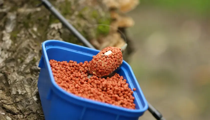 Niebieskie pudełko z przynętą na ryby. W środku mnóstwo pomarańczowych granulek, a na wierzchu feeder z pelletem, gotowy do łowienia.