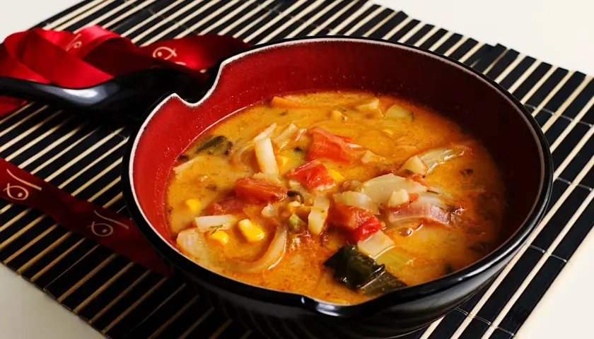 Aromatyczna zupa z surimi z warzywami, podana w misce na macie bambusowej. Obok czarna łyżka i pałeczki ozdobione czerwoną wstążką.