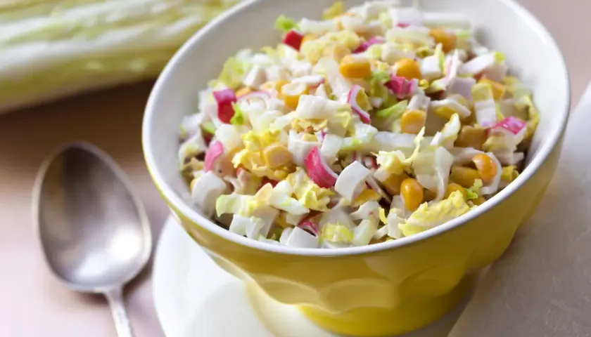 Lekka sałatka z surimi, jajka, kukurydzy i kapusty pekińskiej w żółtej miseczce. Obok łyżka.