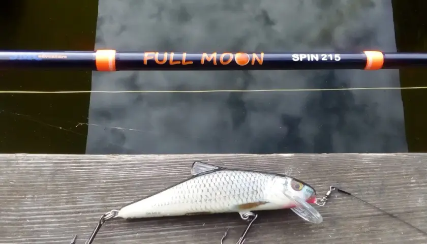 Jaki spinning na szczupaka z brzegu? Wędka "Full Moon Spin 215" i realistyczna przynęta w kształcie ryby czekają na wodzie.