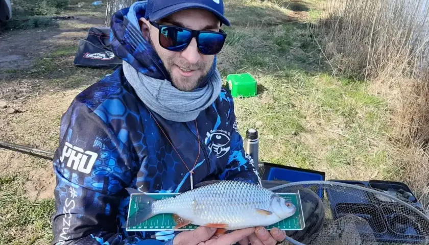 Wędkarz z płocią na miarce, obok zielone pudełko z zanętą na płocie.
