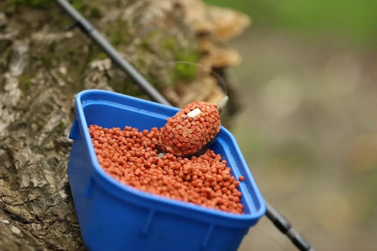 Niebieskie pudełko z przynętą na ryby. W środku mnóstwo pomarańczowych granulek, a na wierzchu feeder z pelletem, gotowy do łowienia.