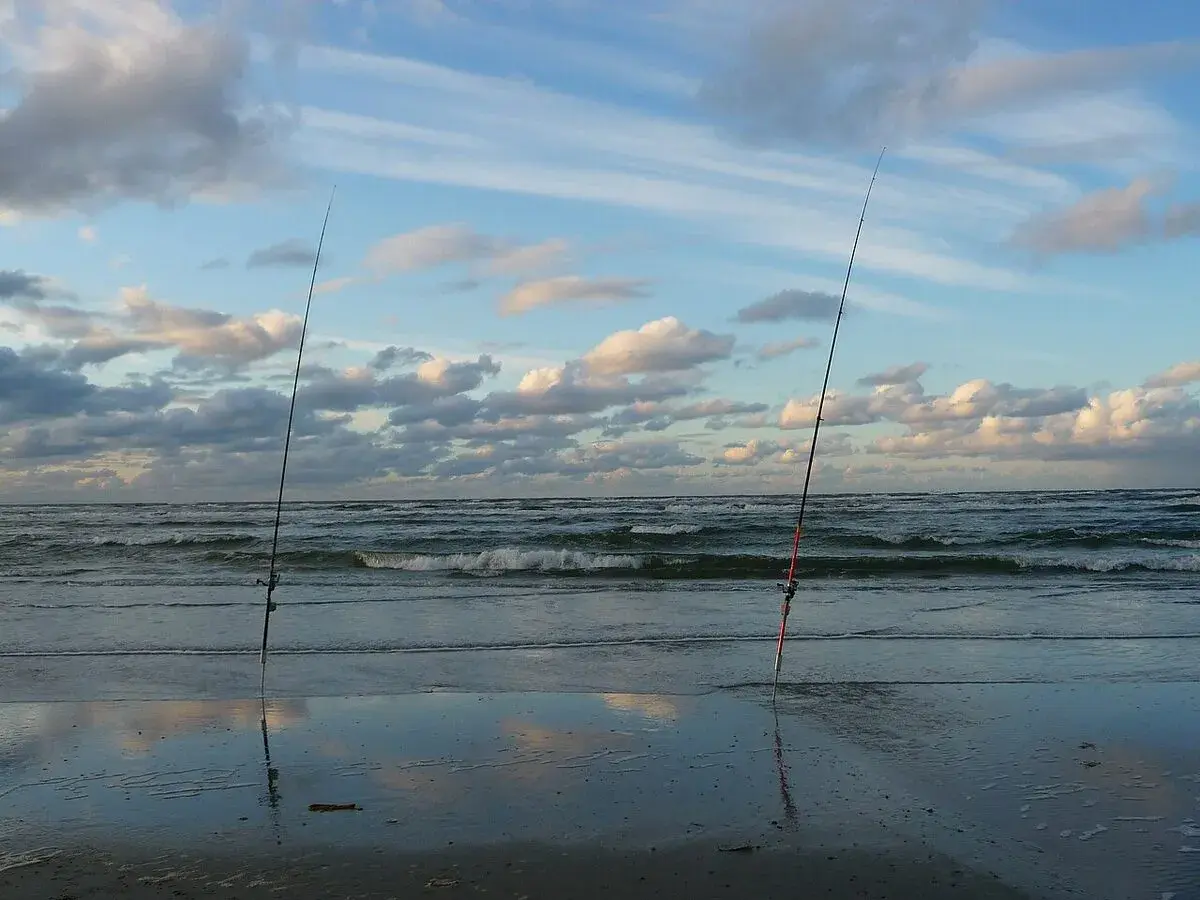 Dwa wędki stoją na plaży, gotowe do trollingu wędkarstwa. Morze faluje pod chmurami.