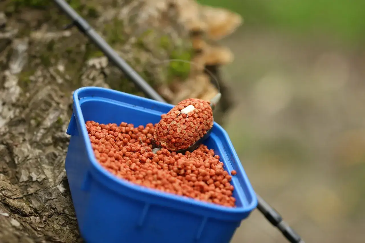 Niebieskie pudełko z przynętą na ryby. W środku mnóstwo pomarańczowych granulek, a na wierzchu feeder z pelletem, gotowy do łowienia.