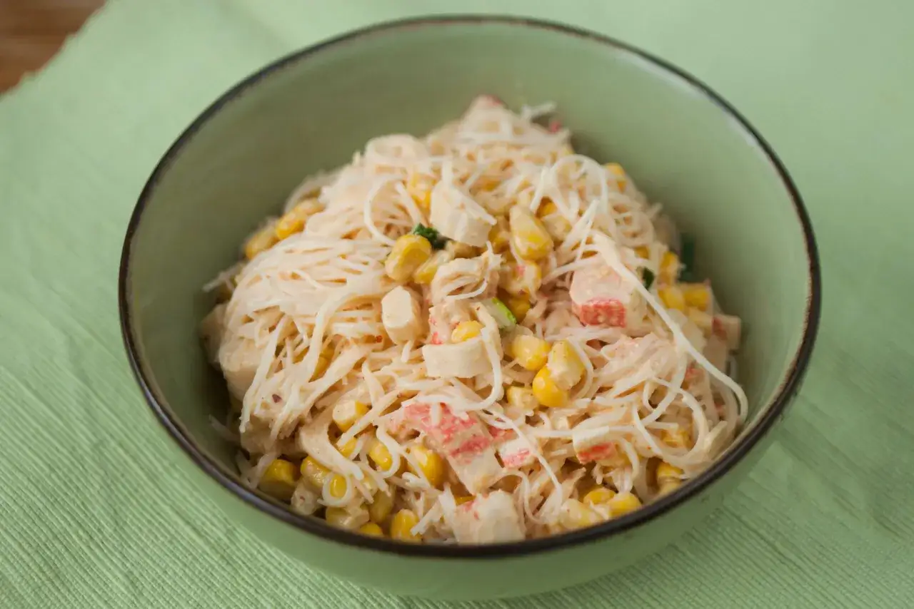 Miseczka z lekką sałatką z surimi, makaronem ryżowym i kukurydzą, podana na zielonym obrusie.