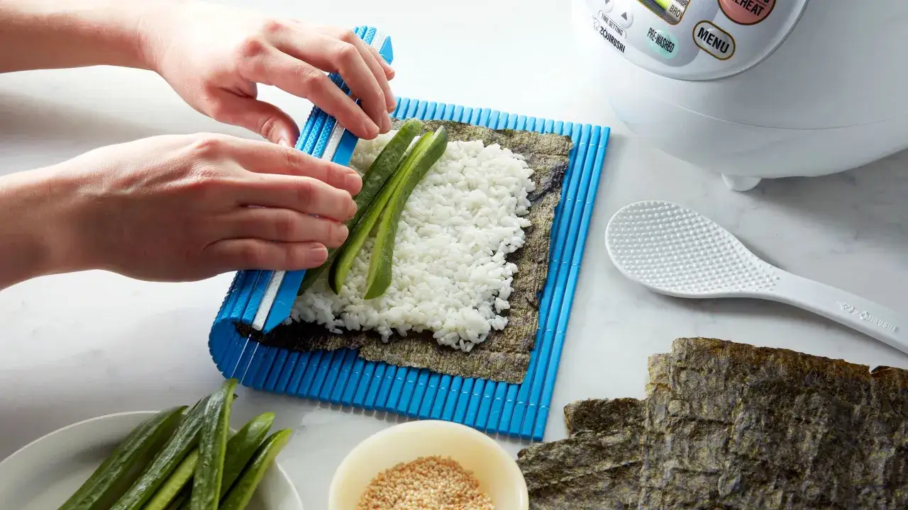 Ręce układają ogórka na ryżu na macie bambusowej, przygotowując się do tego, jak zrobić sushi. Obok leżą płaty nori i sezam.