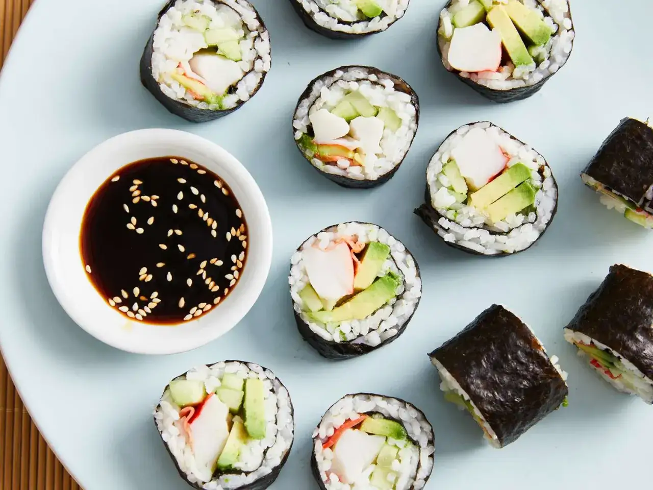 Roladki sushi z awokado, ogórkiem i paluszkami krabowymi, obok miseczka sosu sojowego z sezamem. Idealne składniki sushi na lekki posiłek.