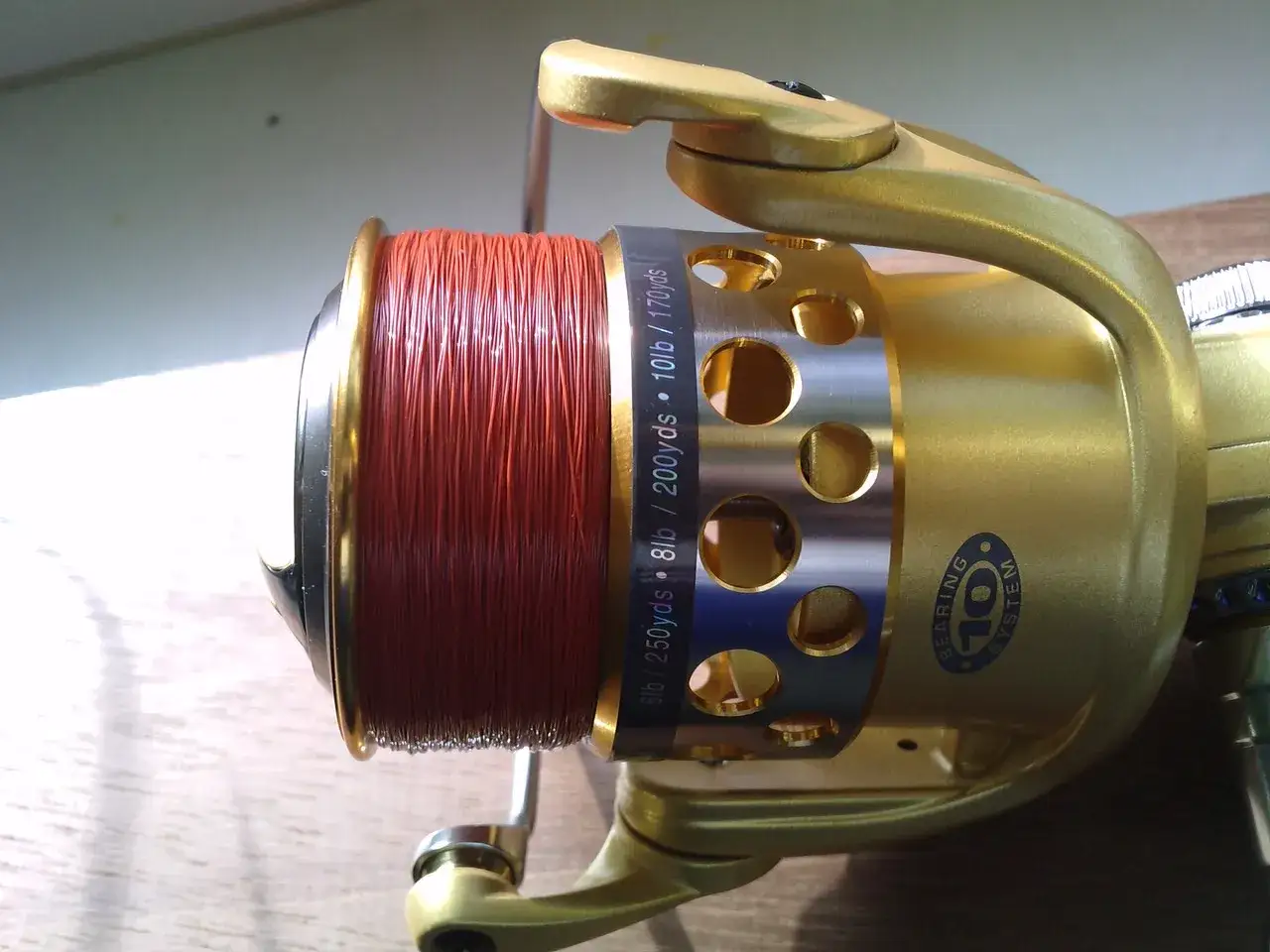 Złoty kołowrotek wędkarski z nawiniętą pomarańczową żyłką. Na obudowie widoczny napis: 6lb / 250yds, 8lb / 200yds, 10lb / 170yds - ile żyłki na kołowrotek.