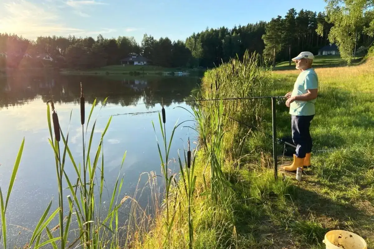 Mężczyzna łowi ryby na jeziorze Kalwa. Słońce odbija się w wodzie, a trzciny szumią na brzegu.