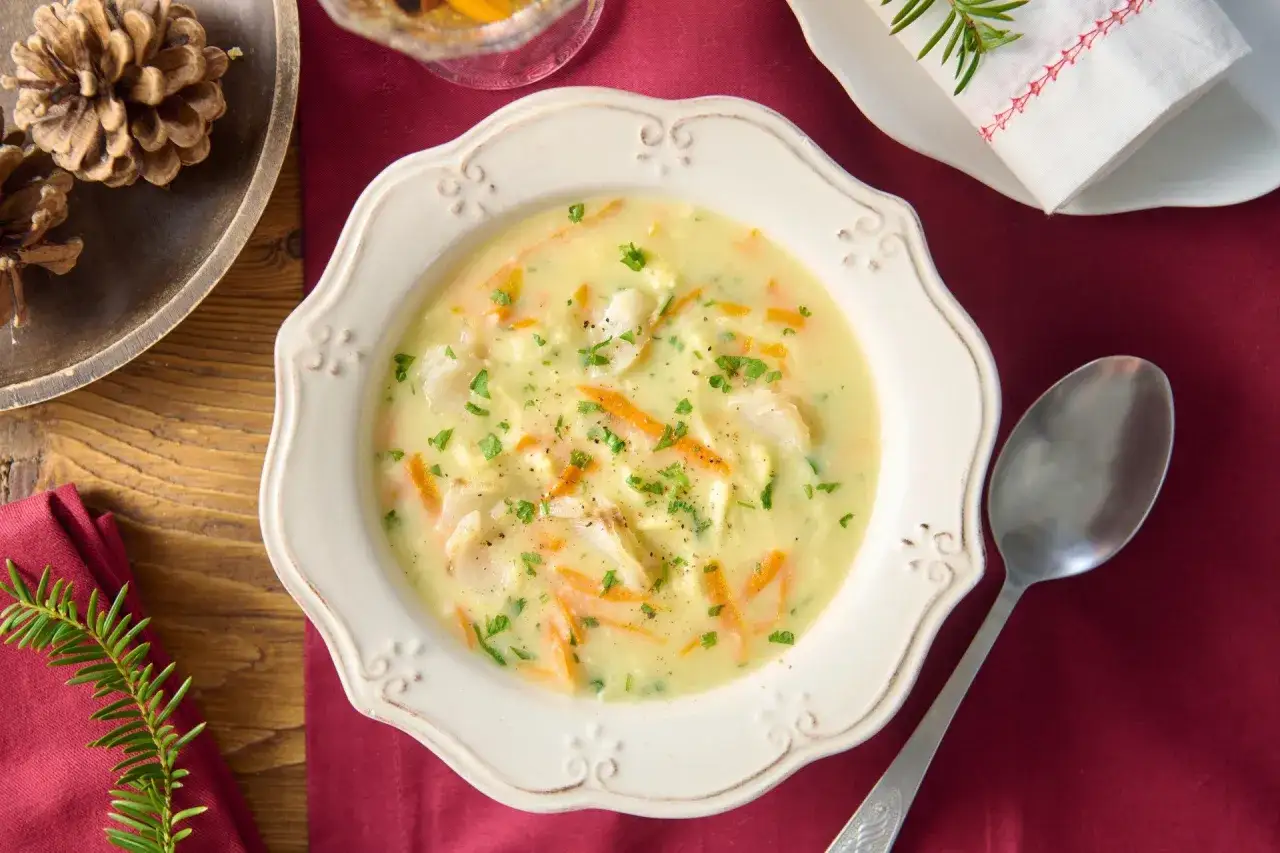 A creamy zupa rybna z kawałkami ryby i marchewki, posypana natką pietruszki. Podana w ozdobnej misce, obok leży łyżka.